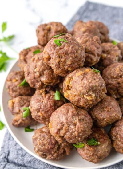 Cooked Parmesan meatballs stacked together on a white plate and topped with fresh chopped parsley.