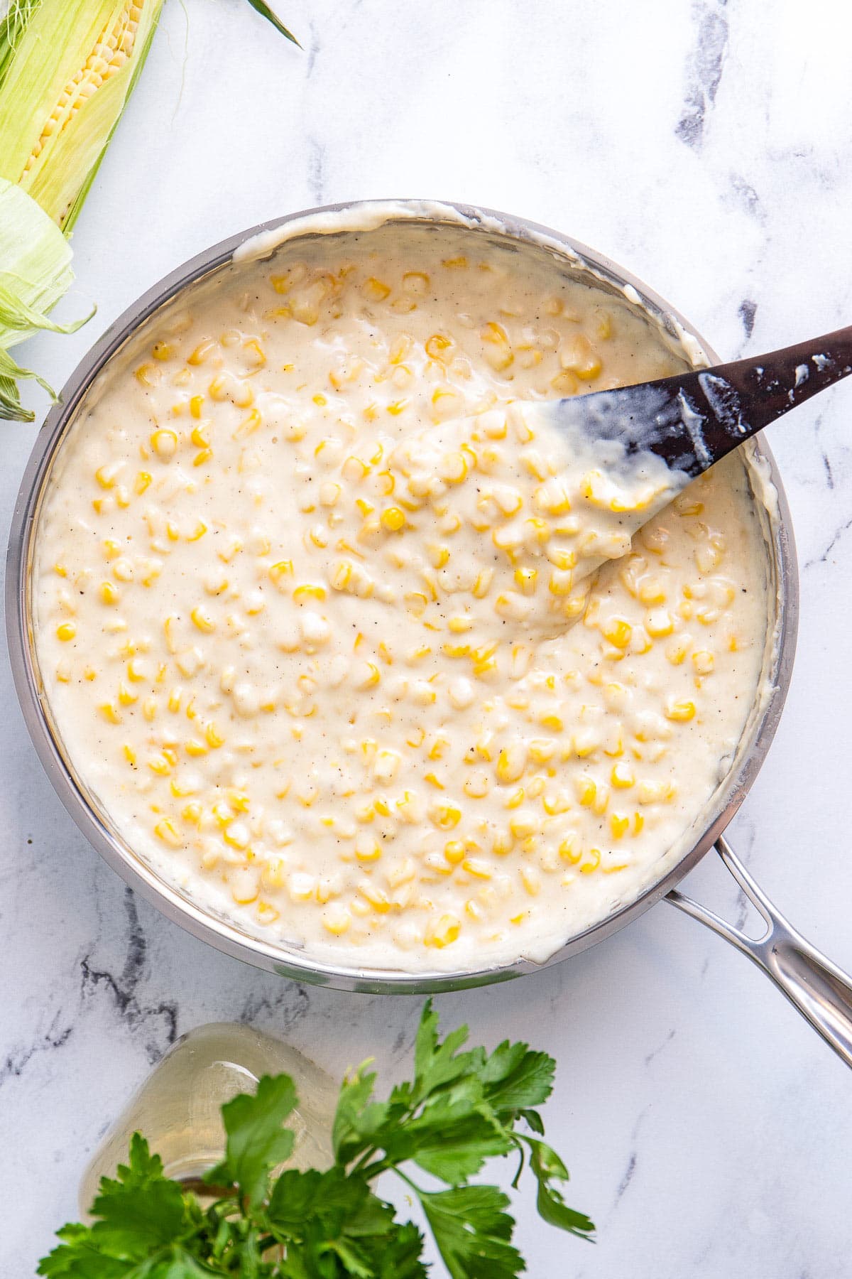 Creamed corn in a stainless steel skillet with corn on the cob and fresh parlsey on the side.