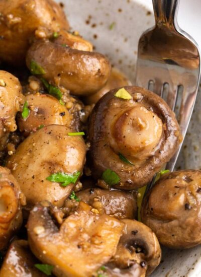 A close up shot of garlic butter mushrooms in a speckled white bowl topped with freshly chopped parsley.