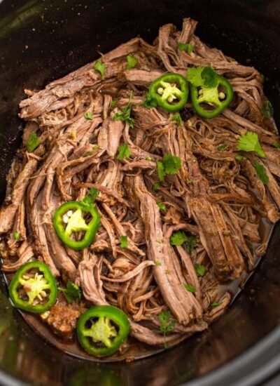 Shredded slow cooker carne asada in a crockpot topped with jalapeno slices and fresh cilantro.