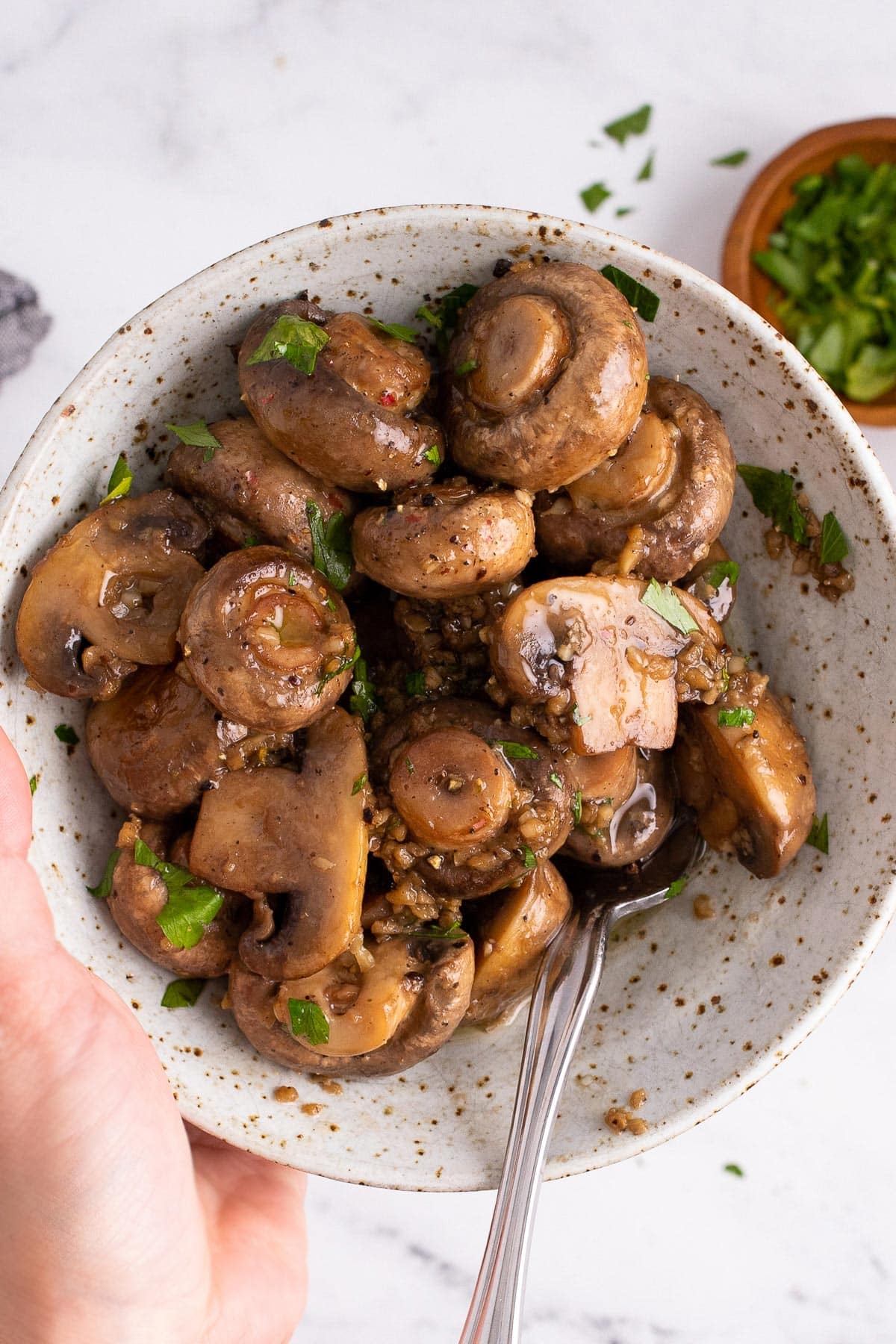 Some garlic butter mushrooms in a speckled white bowl topped with freshly chopped parsley.
