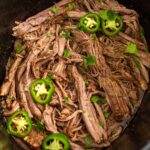 Shredded slow cooker carne asada in a crockpot topped with jalapeno slices and fresh cilantro.