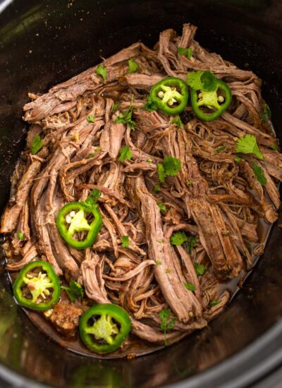 Shredded slow cooker carne asada in a crockpot topped with jalapeno slices and fresh cilantro.