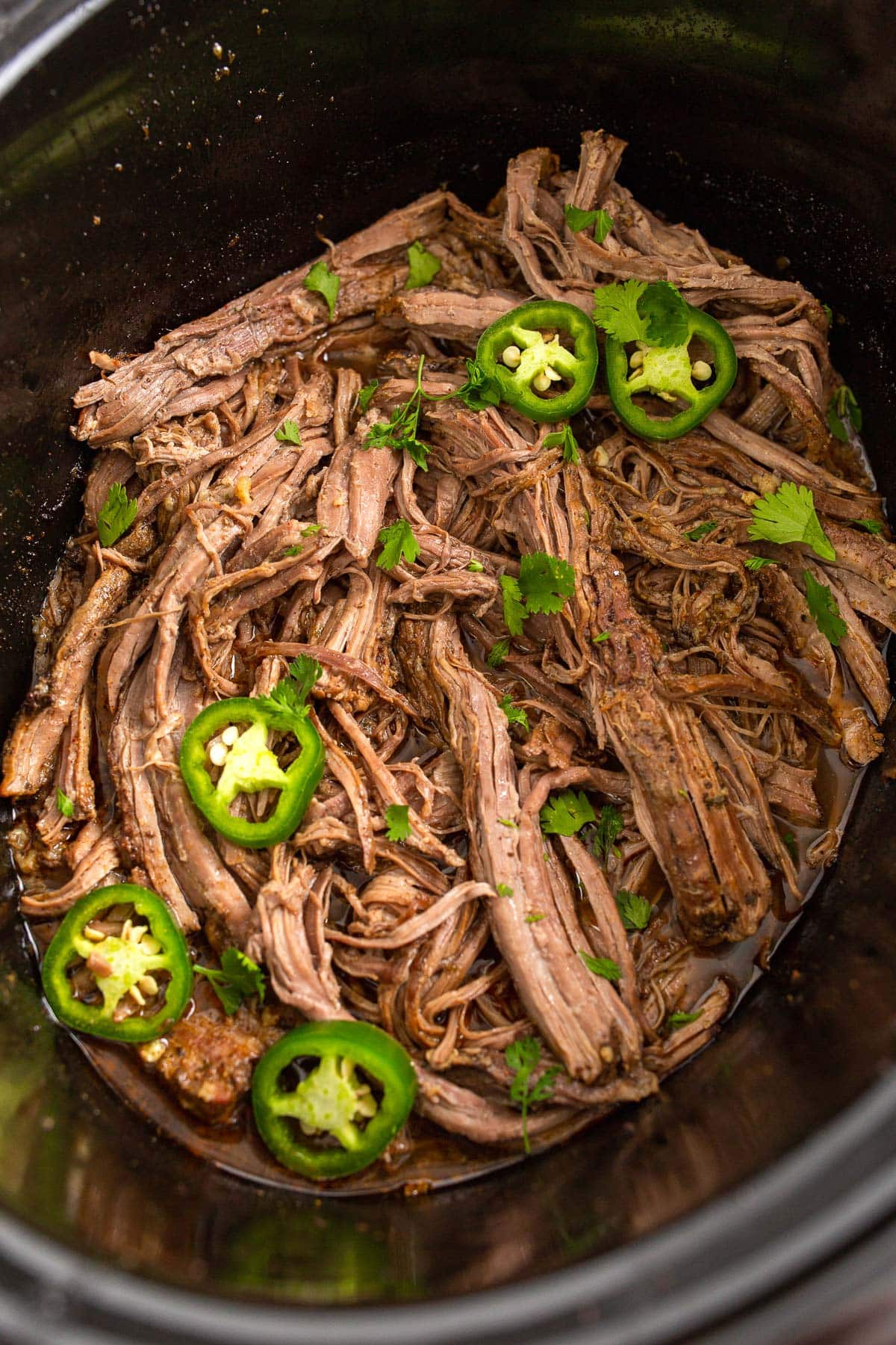 Shredded slow cooker carne asada in a crockpot topped with jalapeno slices and fresh cilantro.