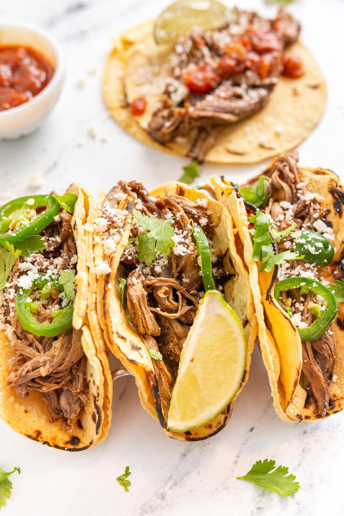 Four corn tortilla stuffed with shredded slow cooker carne asada and topped with jalapeno slices, cilantro and queso fresco.