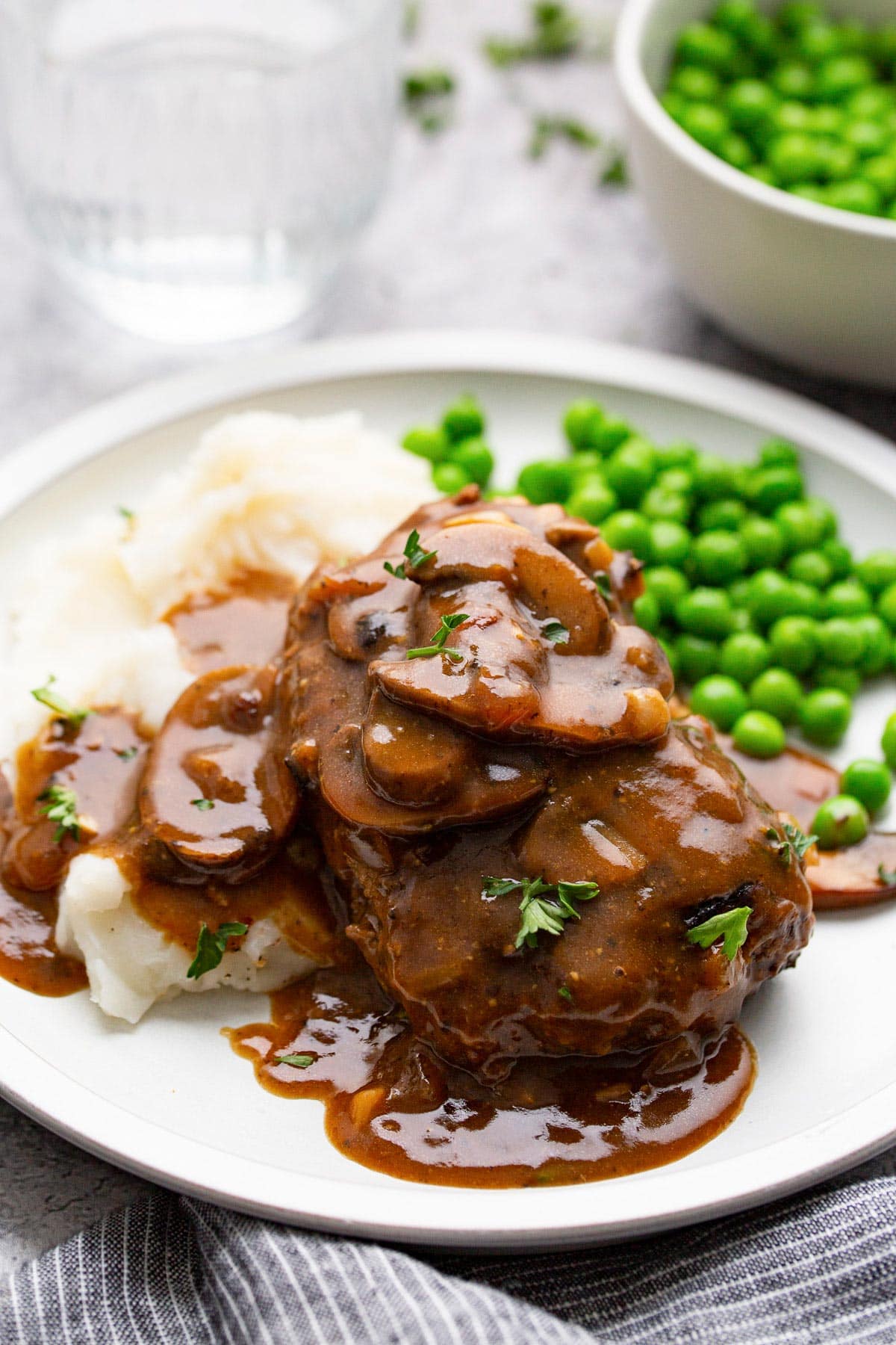Salisbury steak topped with mushroom gravy on a white plate with mashed potatoes and peas.