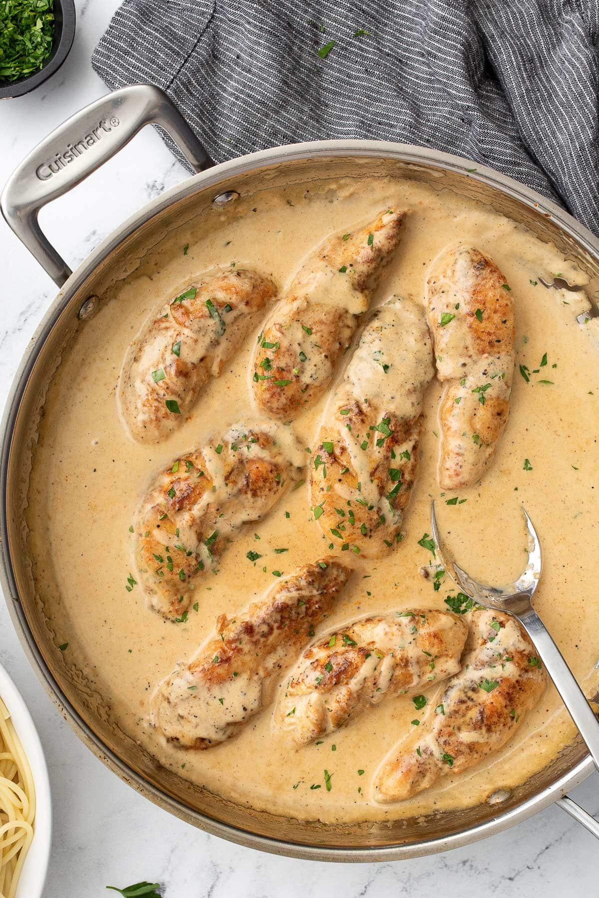 Chicken Lazone in a stainless steel skillet next to a bowl of spaghetti and chopped fresh parsley.