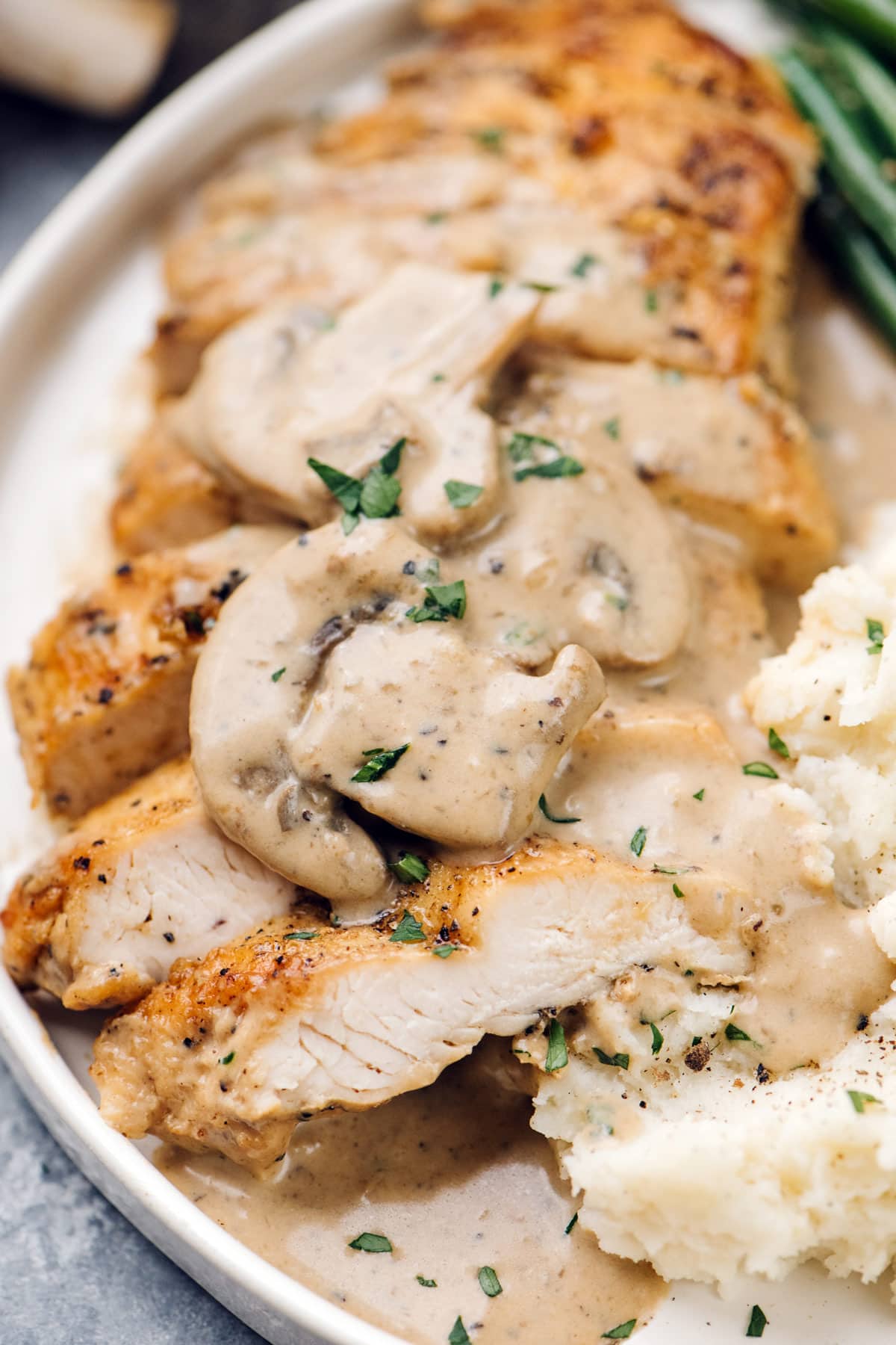 Sliced chicken with creamy mushroom sauce on a cream plate with mashed potatoes and green beans.