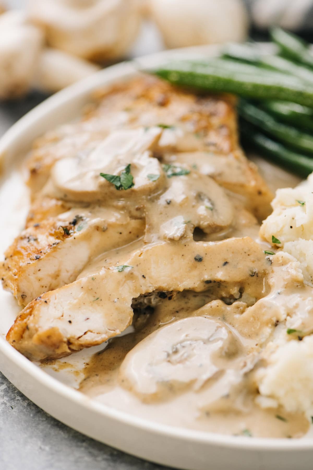 Sliced chicken with creamy mushroom sauce on a cream plate with mashed potatoes and green beans.