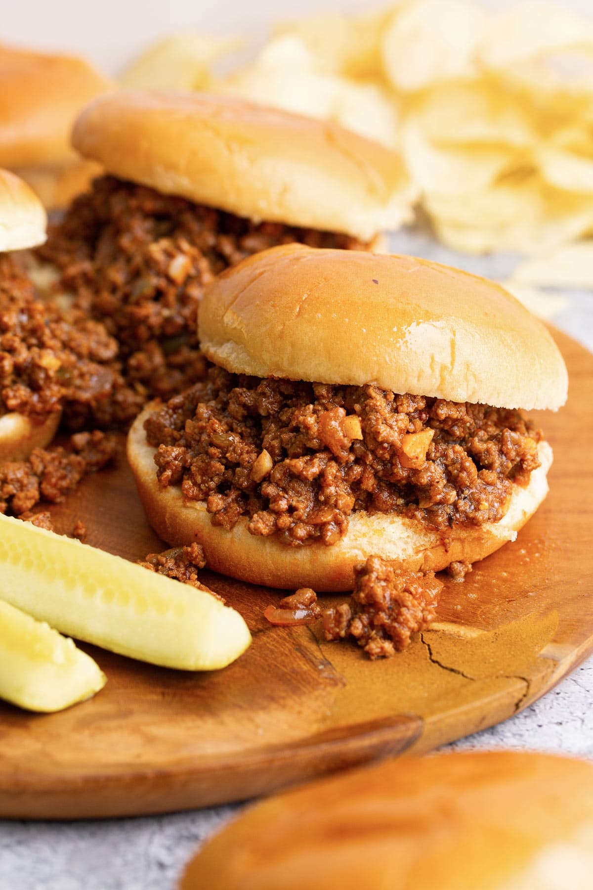 Several homemade Sloppy Joes sandwiches with pickle spears and potato chips on a wooden serving board.