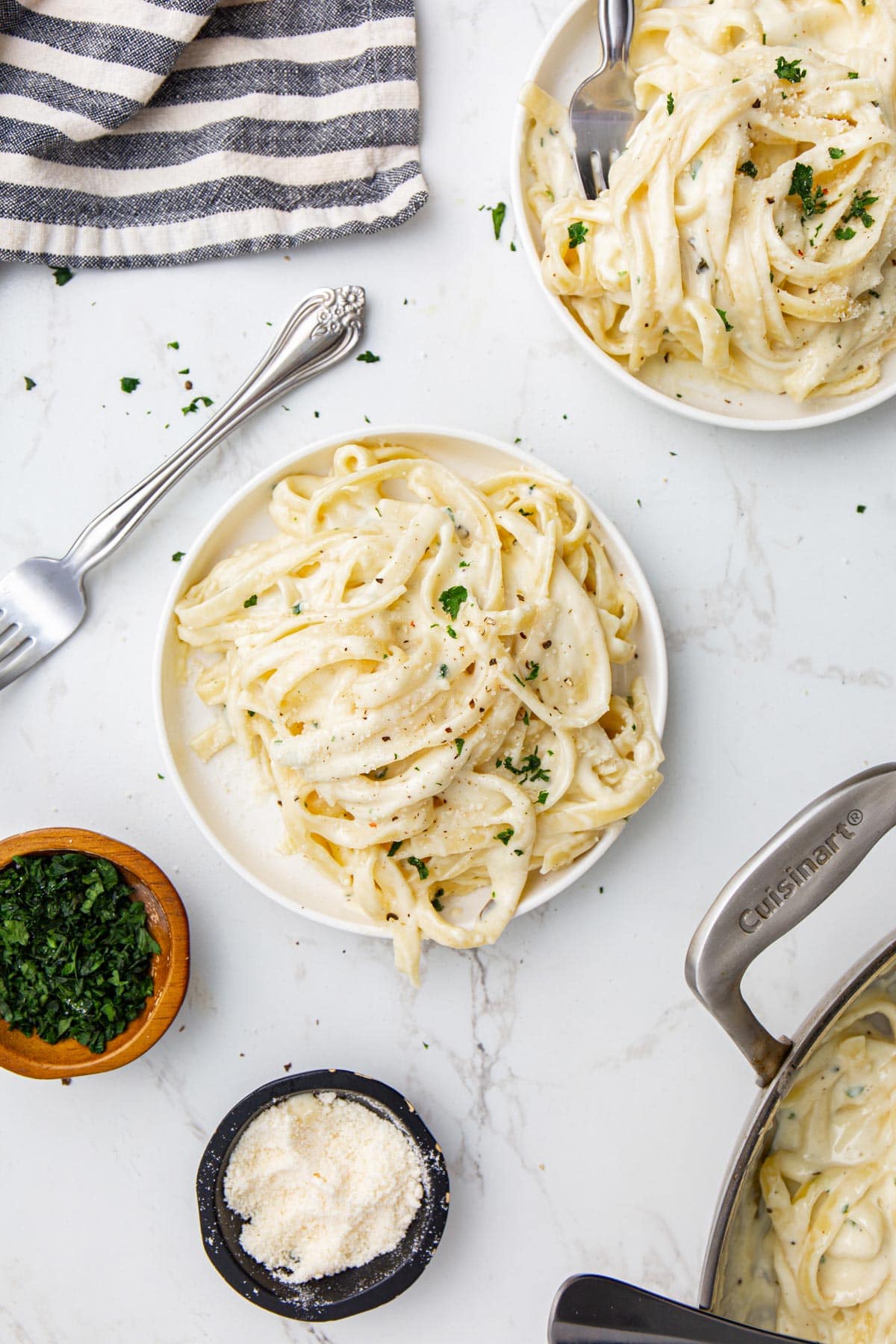 Two servings of homemade Alfredo sauce paired with fettucine pasta on white plates topped with chopped fresh parsley.
