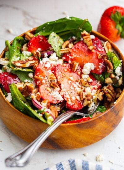 A serving of spinach strawberry salad in a wooden bowl topped with feta cheese and pecans.