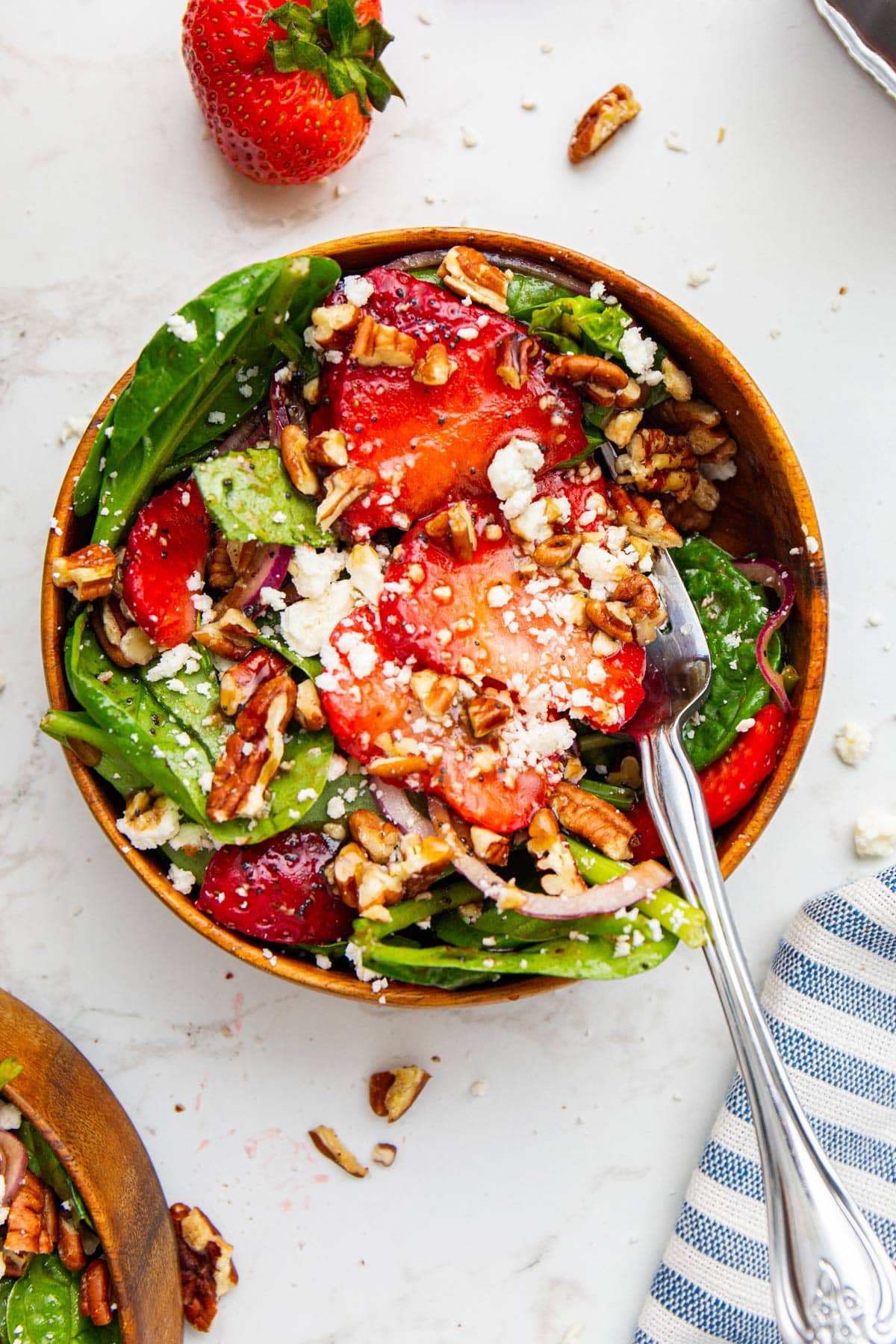 A serving of spinach strawberry salad in a wooden bowl topped with feta cheese and pecans.