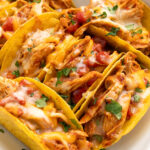 Several cheesy baked chicken tacos stacked together in a white casserole dish topped with chopped fresh cilantro.