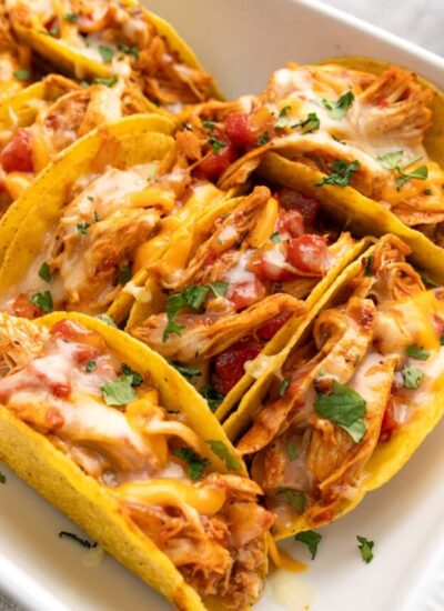 Several cheesy baked chicken tacos stacked together in a white casserole dish topped with chopped fresh cilantro.