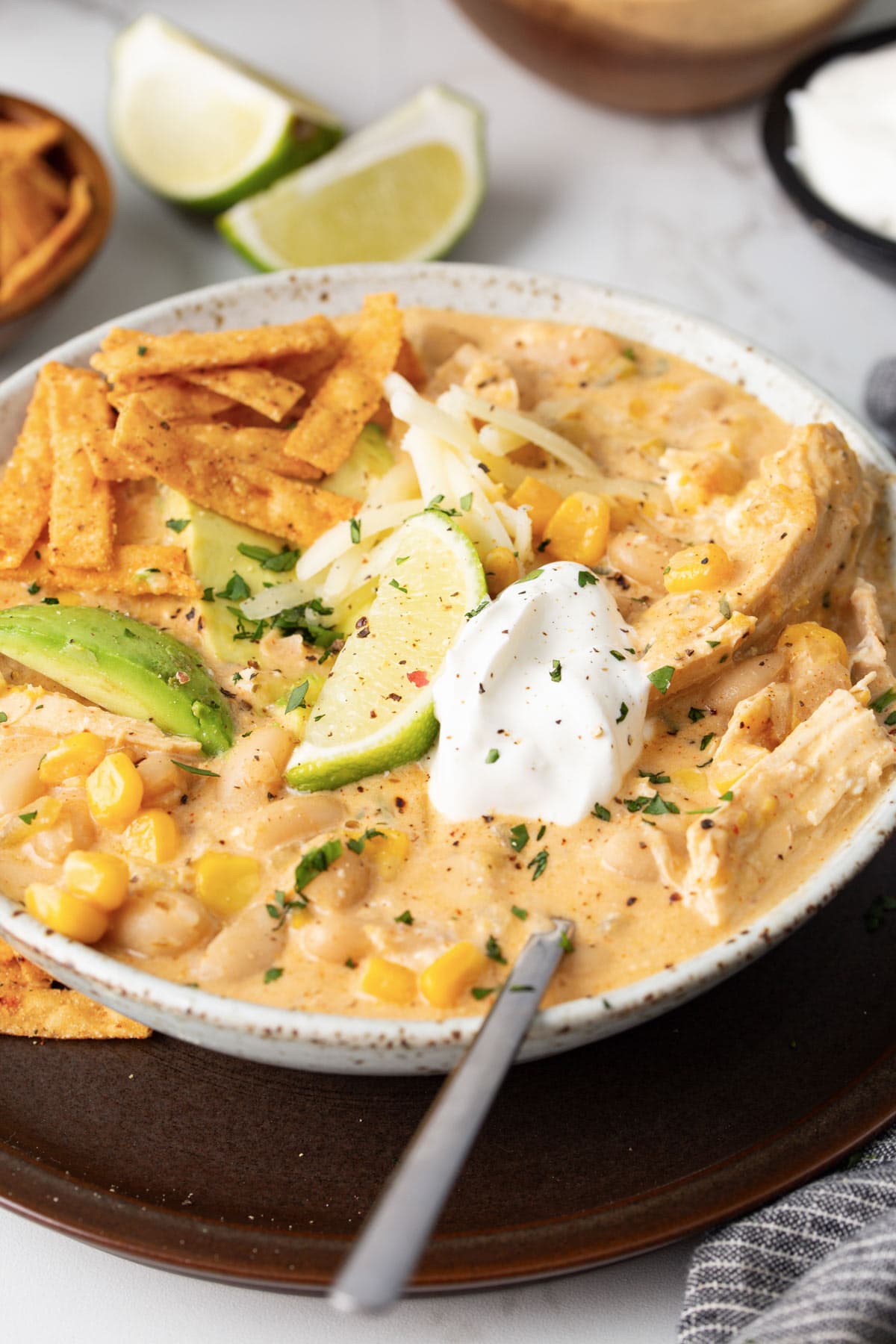 A big bowl of white chicken chili topped with tortilla strips, shredded cheese, sliced avocado, sour cream, lime wedges and chopped cilantro.