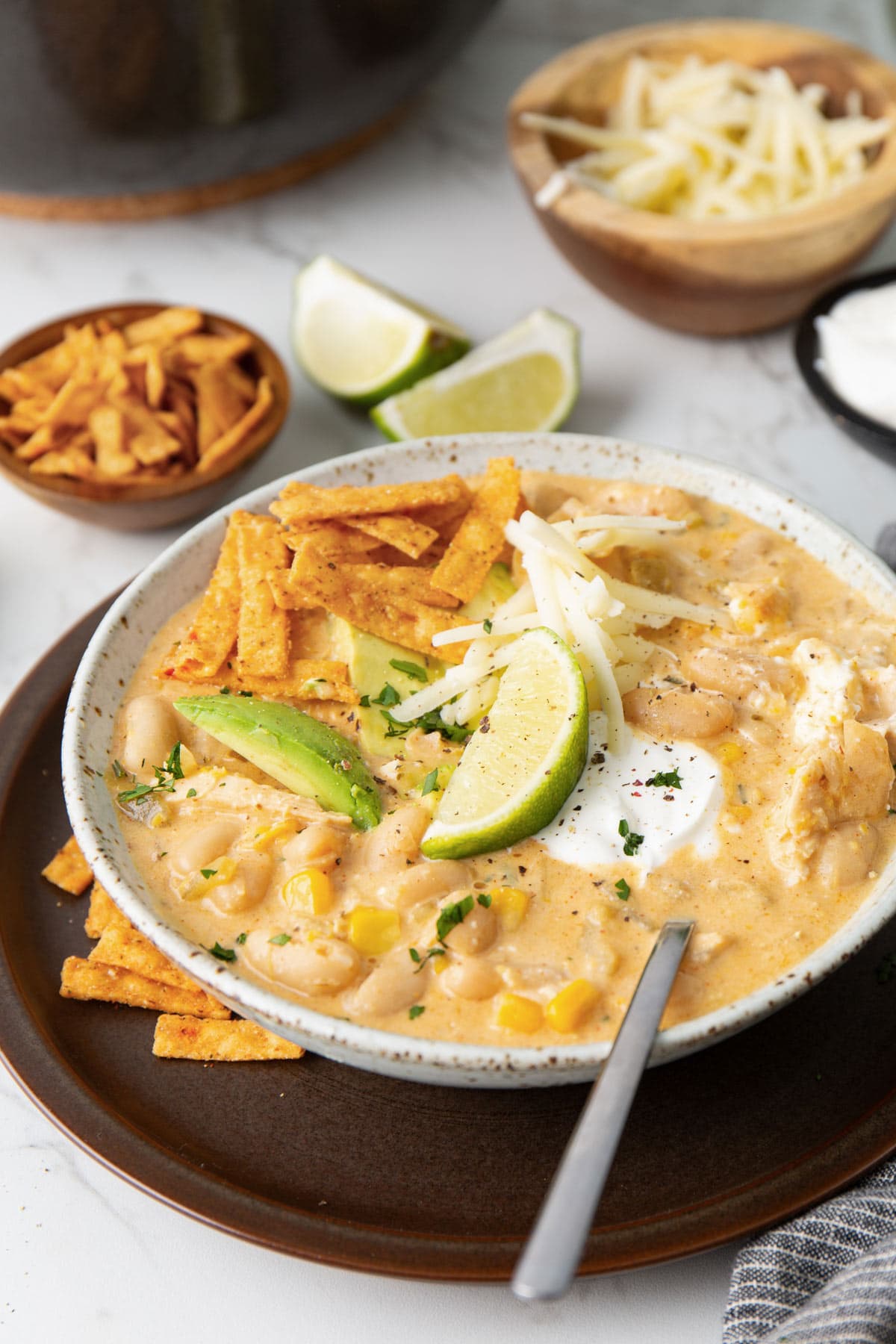 A big bowl of white chicken chili topped with tortilla strips, shredded cheese, sliced avocado, sour cream, lime wedges and chopped cilantro.