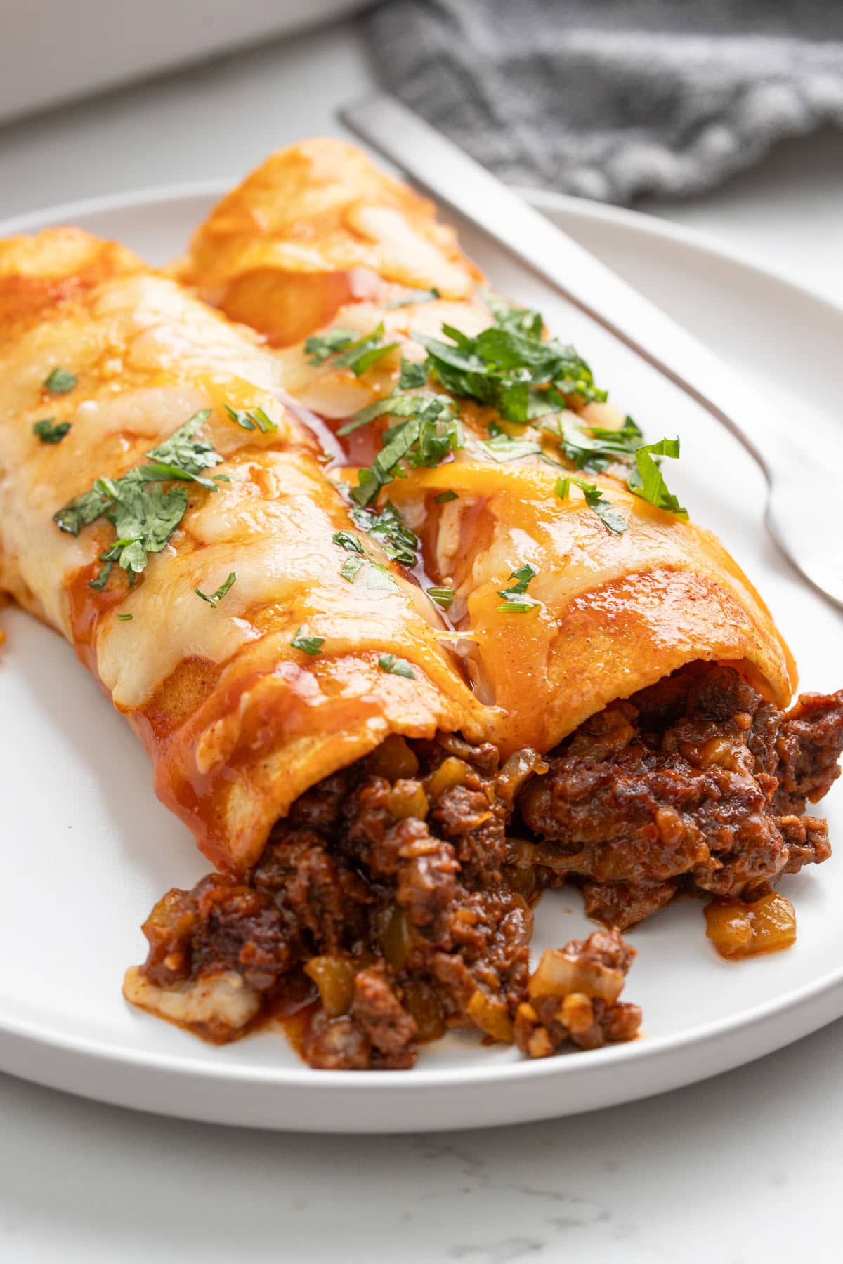 Two ground beef enchiladas on a white plate topped with enchilada sauce, melted cheese and chopped fresh parsley.