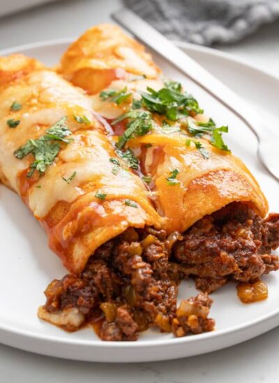 Two ground beef enchiladas on a white plate topped with enchilada sauce, melted cheese and chopped fresh parsley.