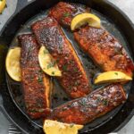 Honey glazed salmon fillets topped with lemon wedges and chopped parsley in a cast iron pan.