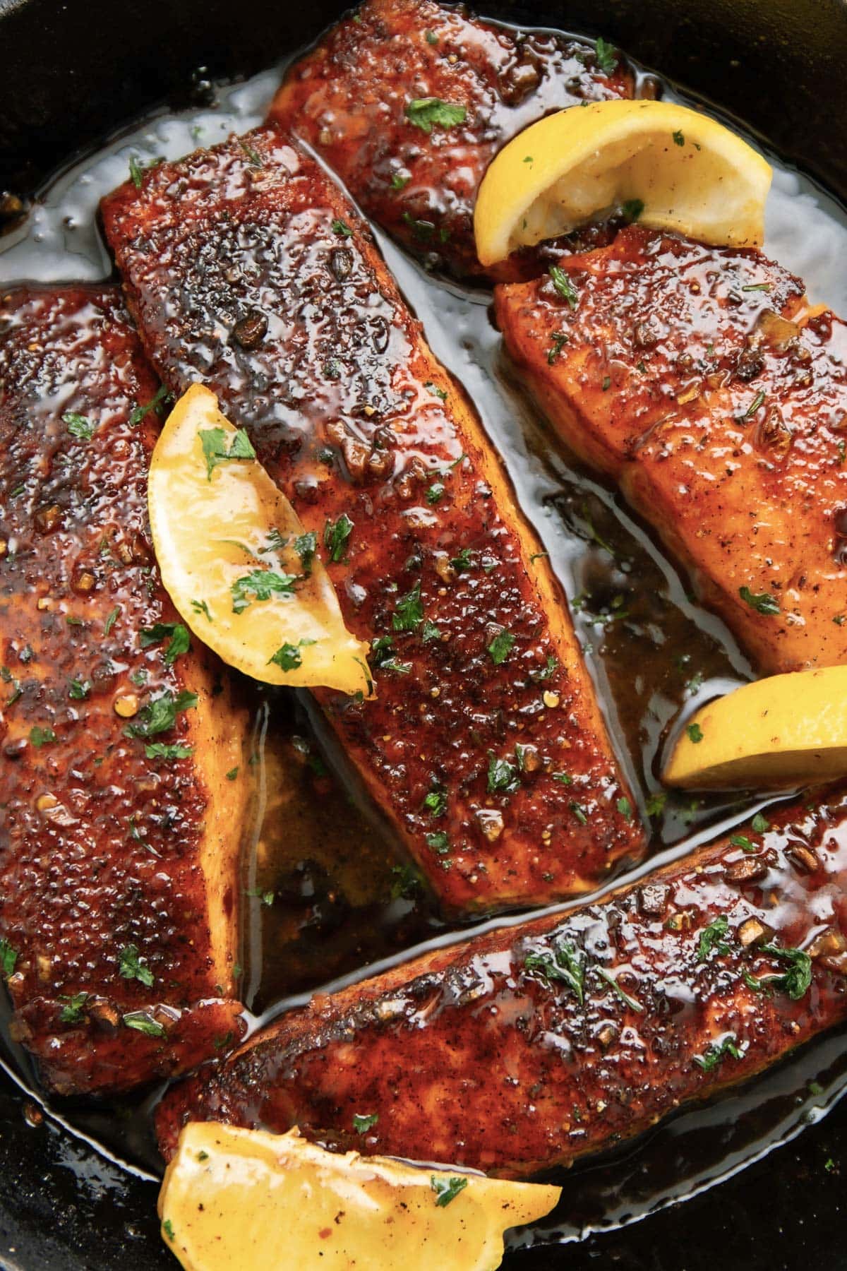 A close up of four honey glazed salmon fillets topped with lemon wedges and chopped parsley in a cast iron pan.