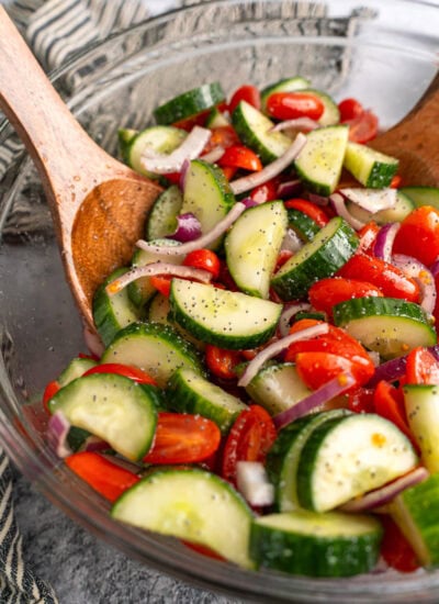 Cucumber tomato salad in a glass mixing bowl tossed with two wooden salad servers.