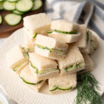 Several cucumber sandwiches stacked high on a white plate with a garnish of fresh dill.