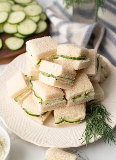 Several cucumber sandwiches stacked high on a white plate with a garnish of fresh dill.