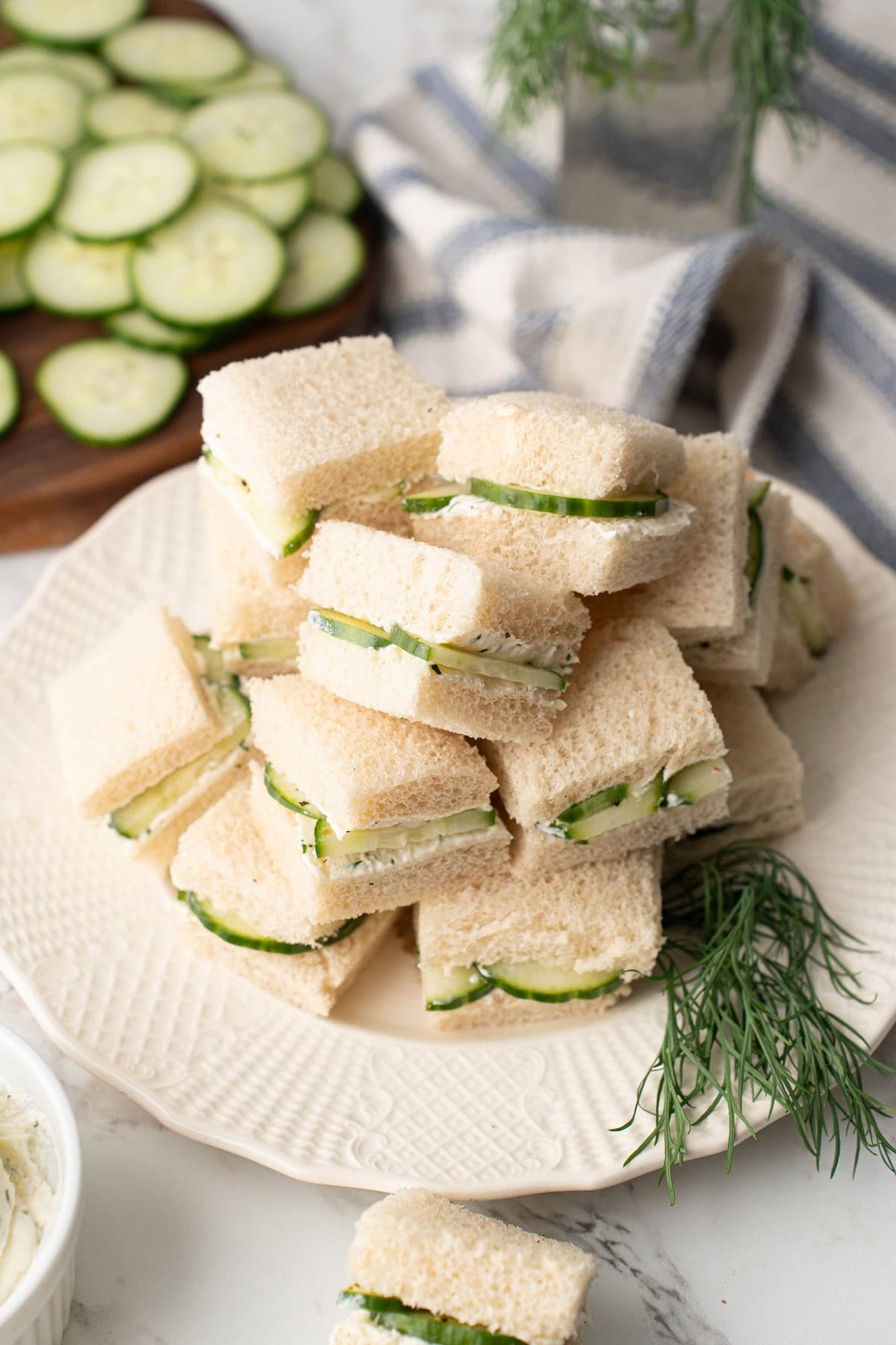 Several cucumber sandwiches stacked high on a white plate with a garnish of fresh dill.