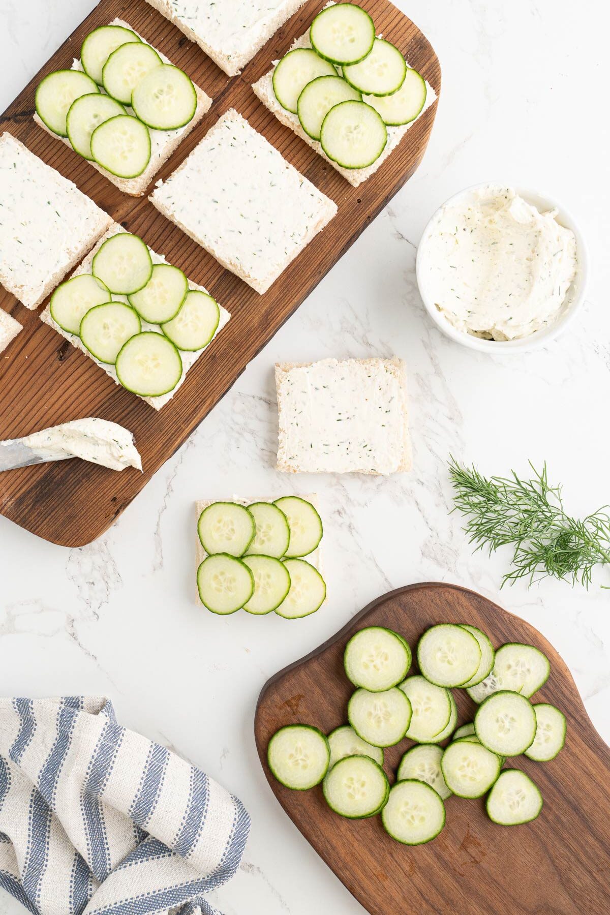 The process of making cucumber sandwiches with crustless bread slathered in cream cheese spread and thinly sliced cucumbers.